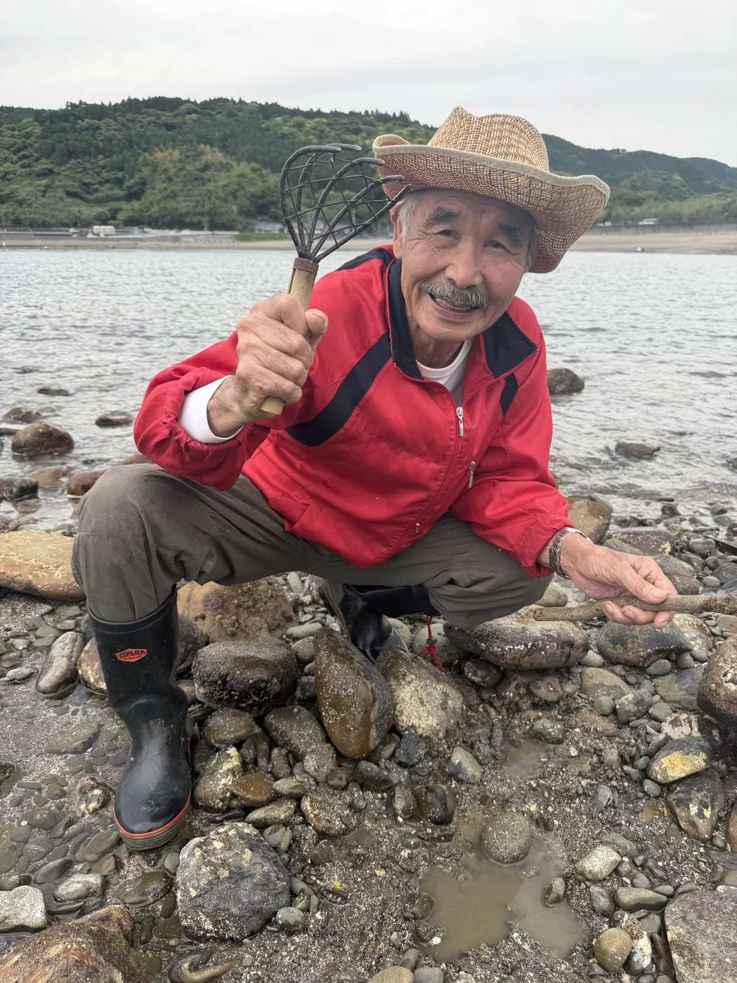 大潮の干潮で潮が引いた浜で、熊手を持ってしゃがむ遠矢国利名人